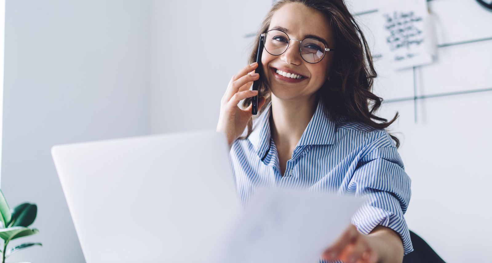 Woman with glasses smiling, on phone, holding paper near laptop in bright office.