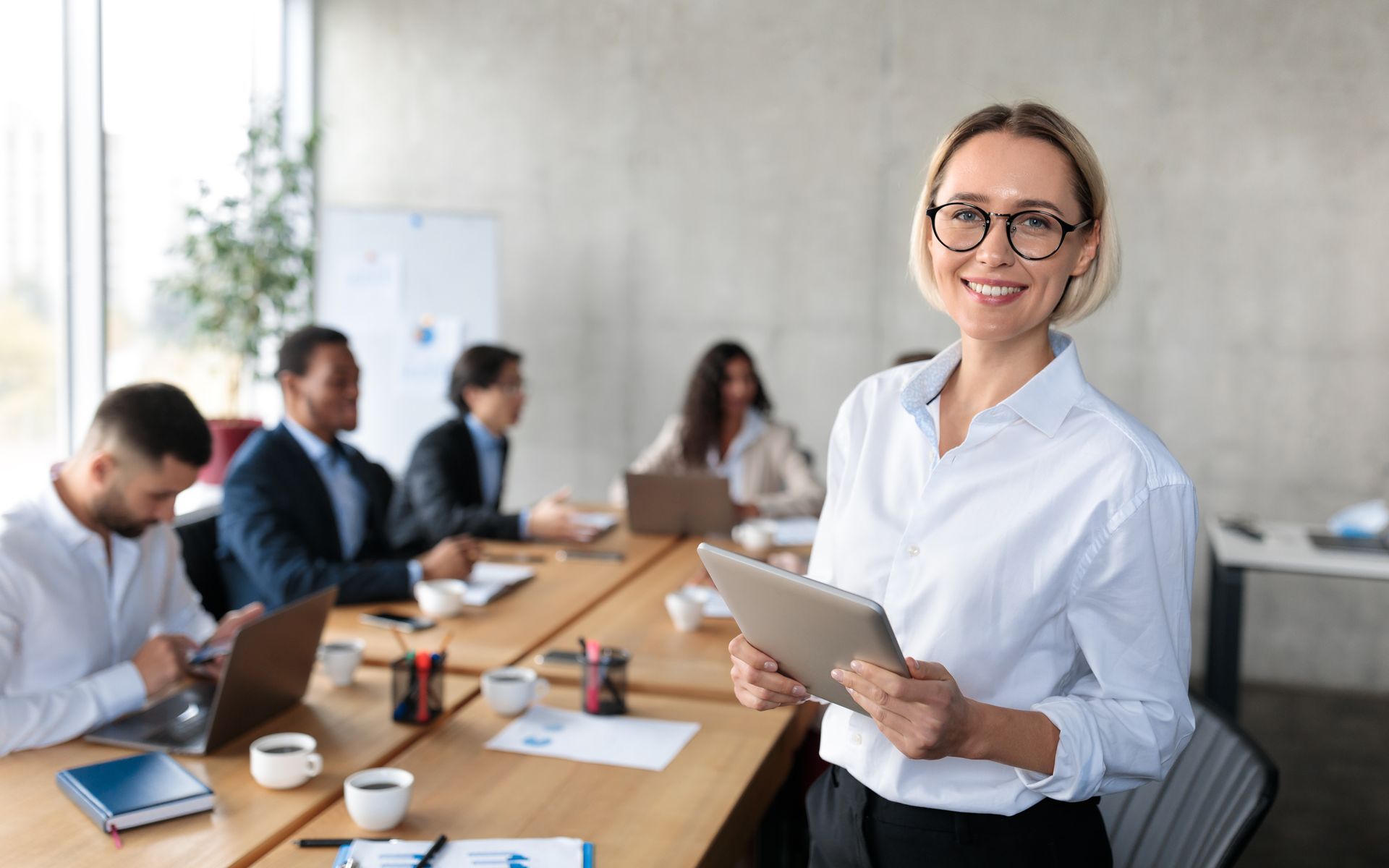 Woman in glasses smiles, holding tablet, at conference table with colleagues.