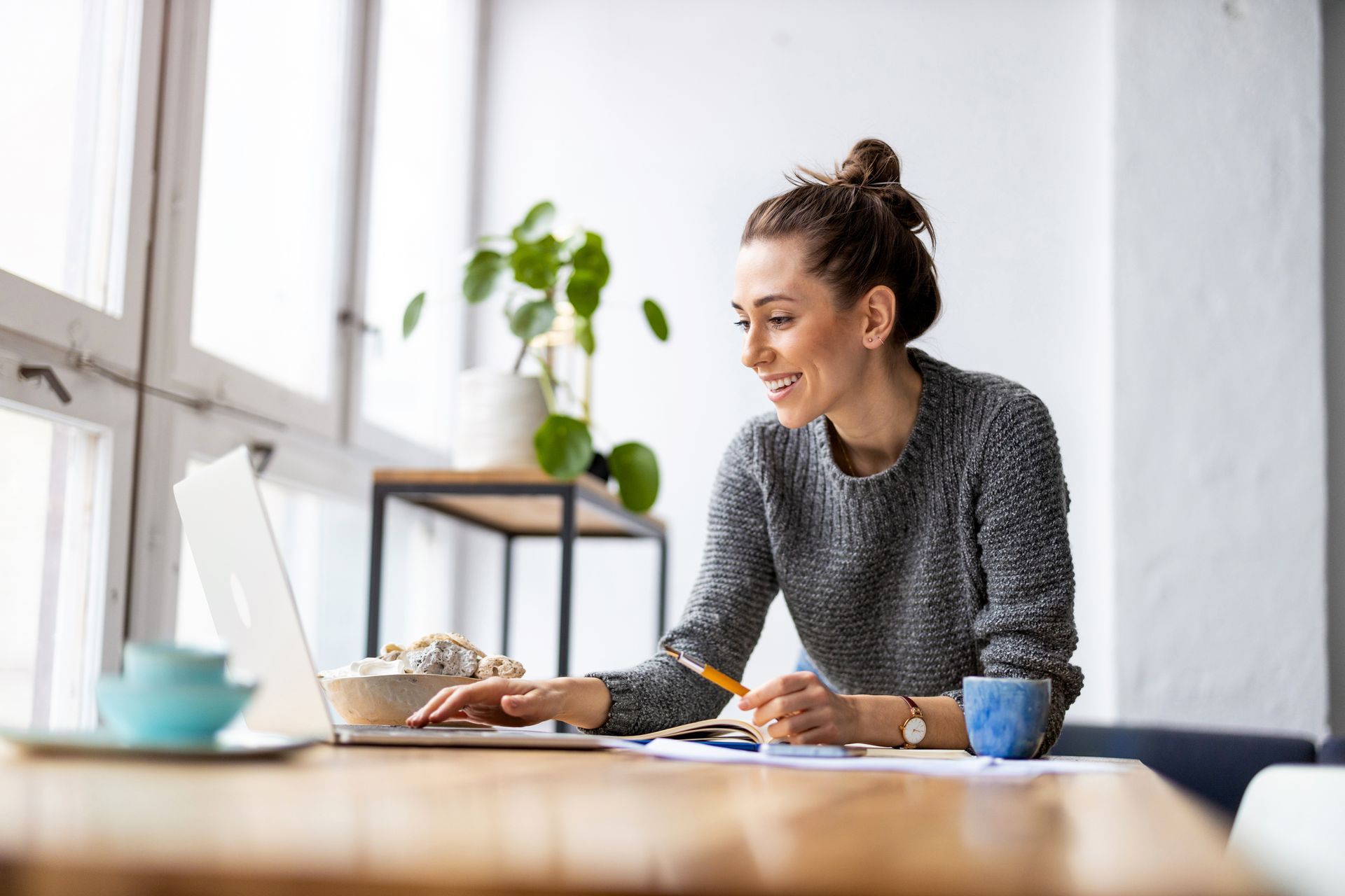 Woman in gray sweater works on laptop at a wooden table near window, smiling and holding a pen.