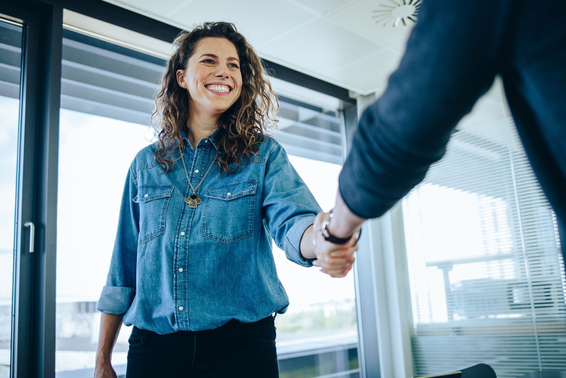 Woman in denim shirt smiling, shaking hands with unseen person in office setting.