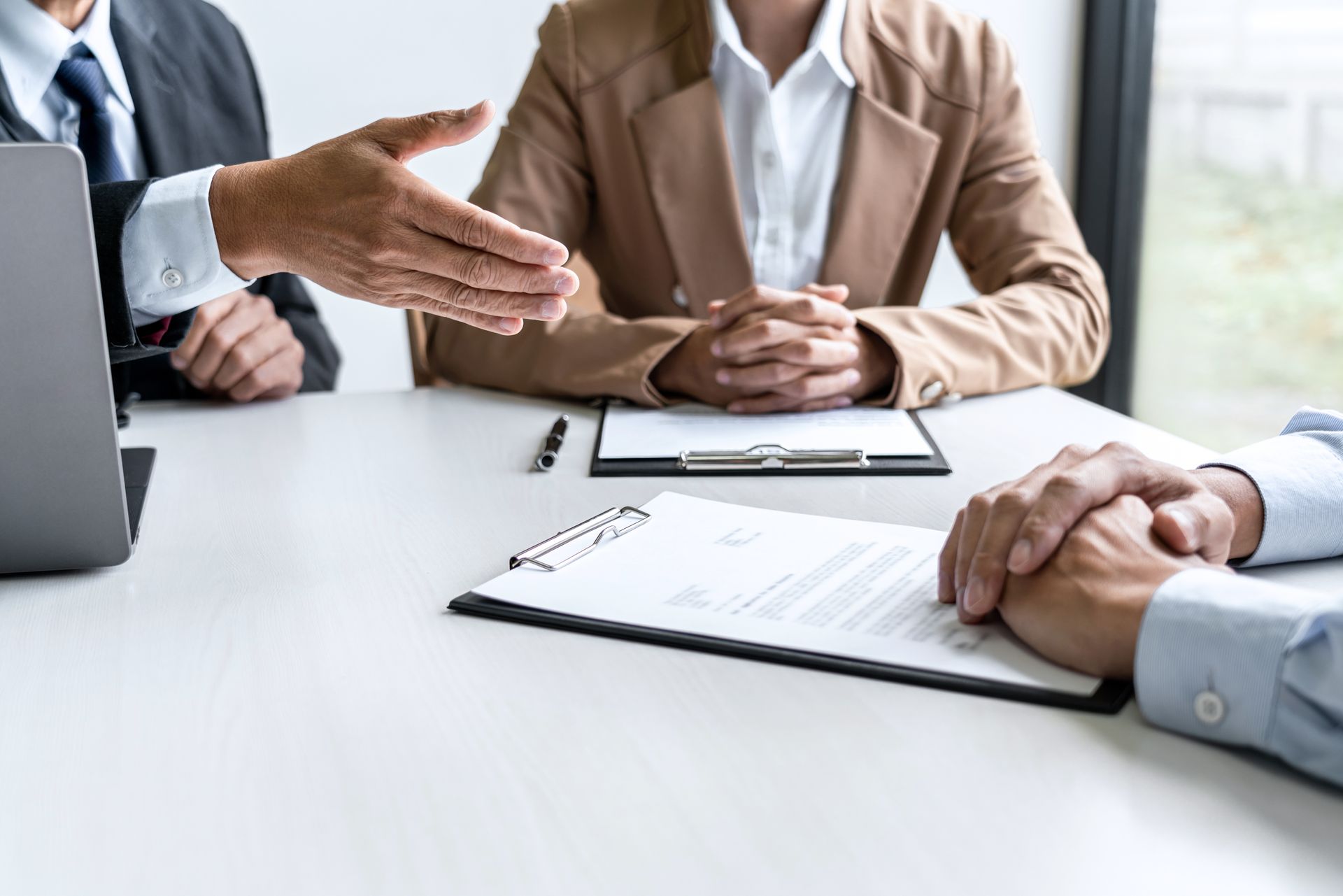 People in business attire at a table; one extends a hand for a handshake, with documents and a laptop visible.