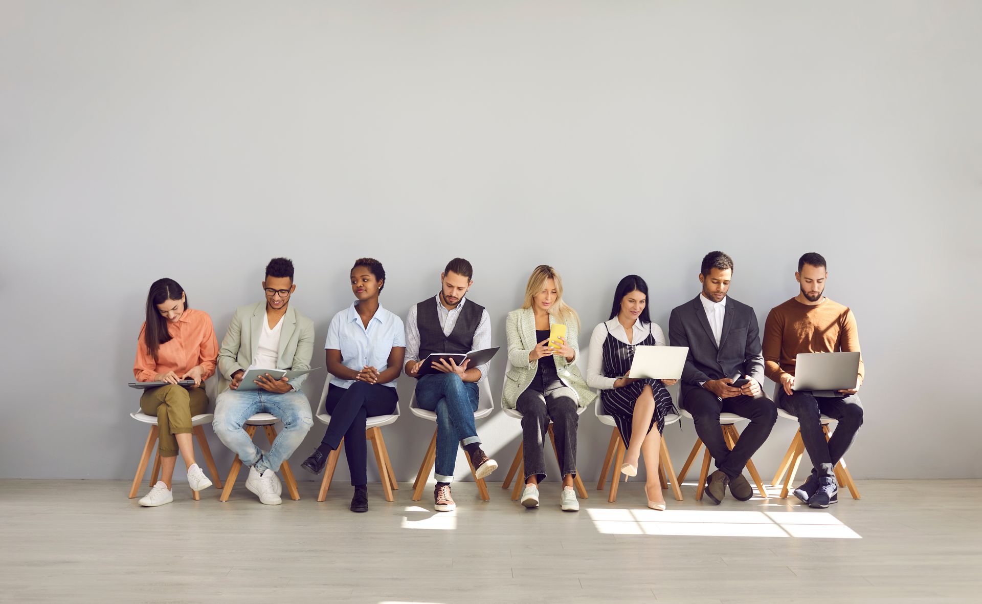People seated in a row, using phones and laptops, against a gray wall.