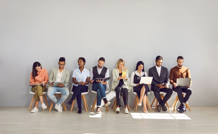 Group of people seated on chairs against a gray wall, some using phones and laptops.