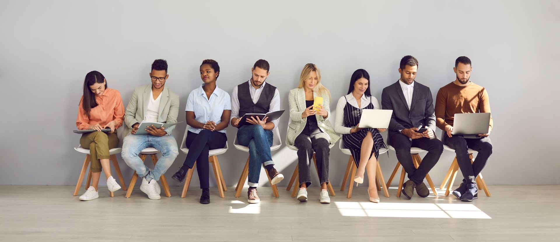 People seated in a row, each with a laptop, tablet, or phone, in a waiting area.