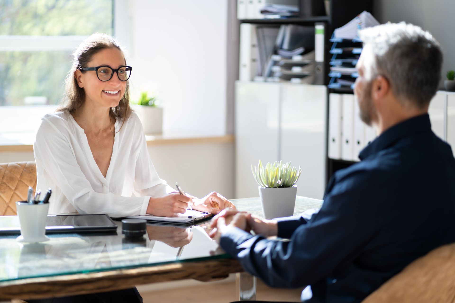Woman in glasses interviewing a man, seated across a desk, in an office setting.