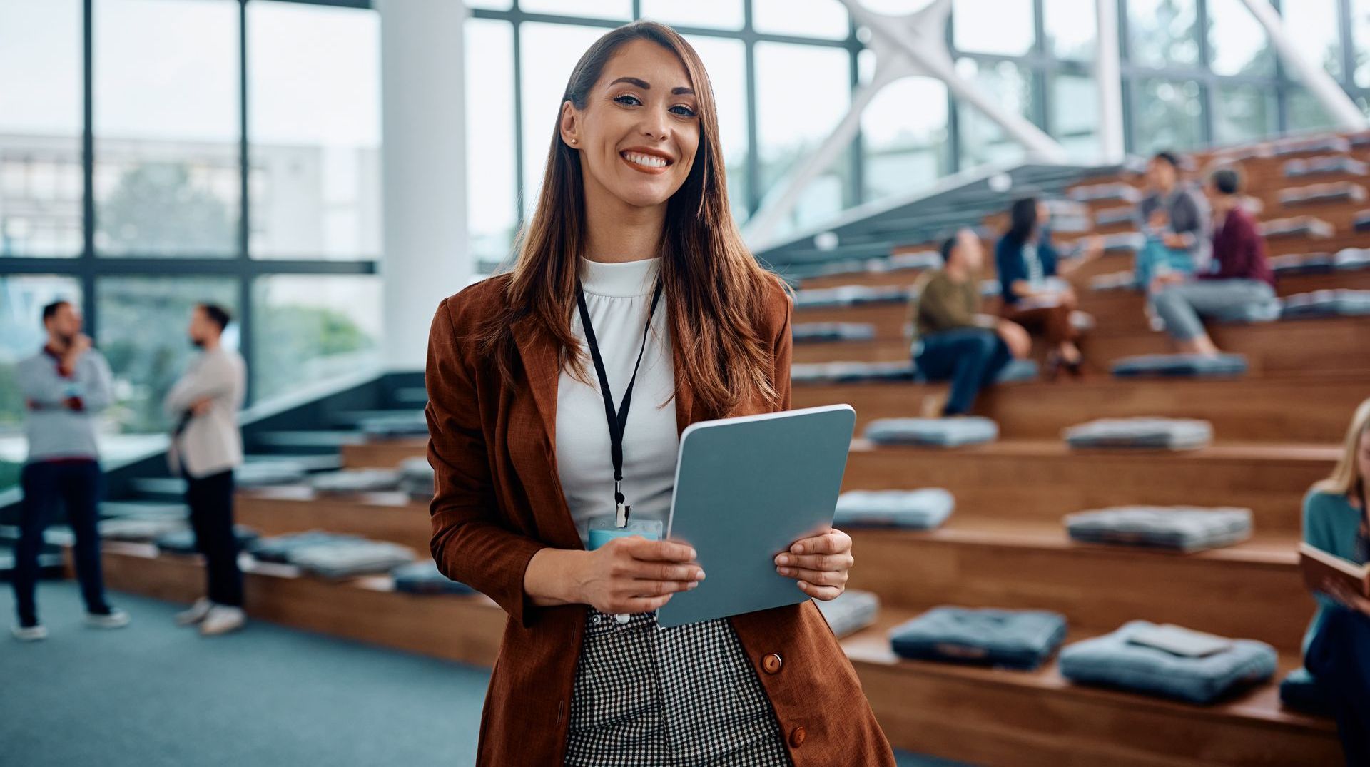 Woman in brown blazer, holding a tablet, smiles at the camera in a modern building with a wooden seating area.