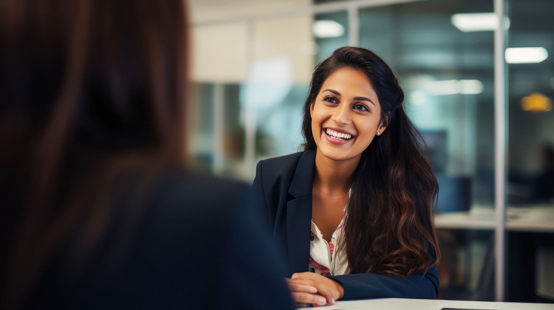 Woman in a blazer smiles and talks to someone off-screen in a bright office.