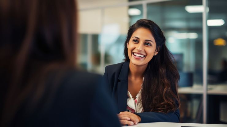 Woman in a blazer smiles and looks at another person in a business setting.