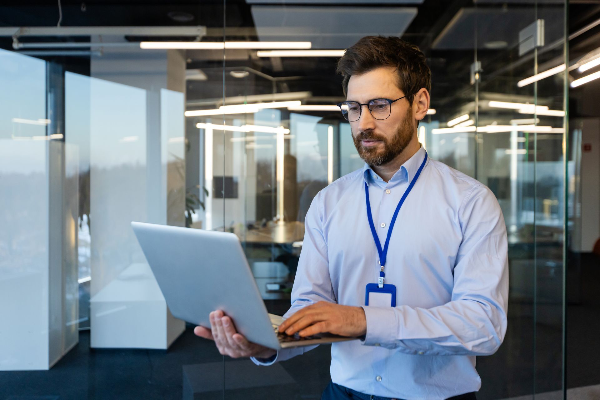 Man wearing glasses, working on a laptop in a modern office with glass walls.