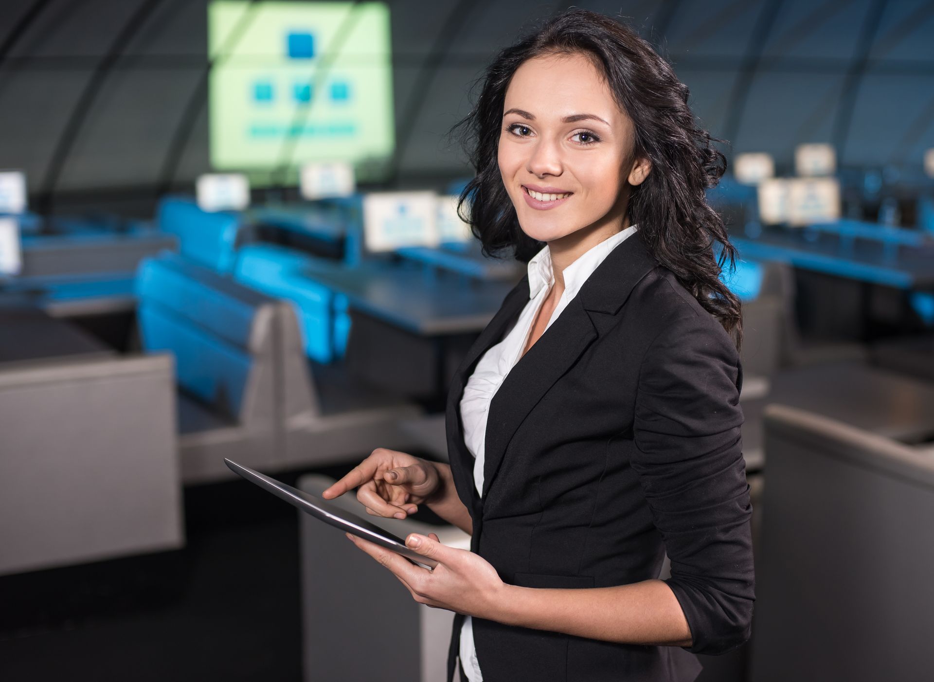 Woman in a blazer smiles, holding a tablet, standing in an office environment with desks and a screen in the background.