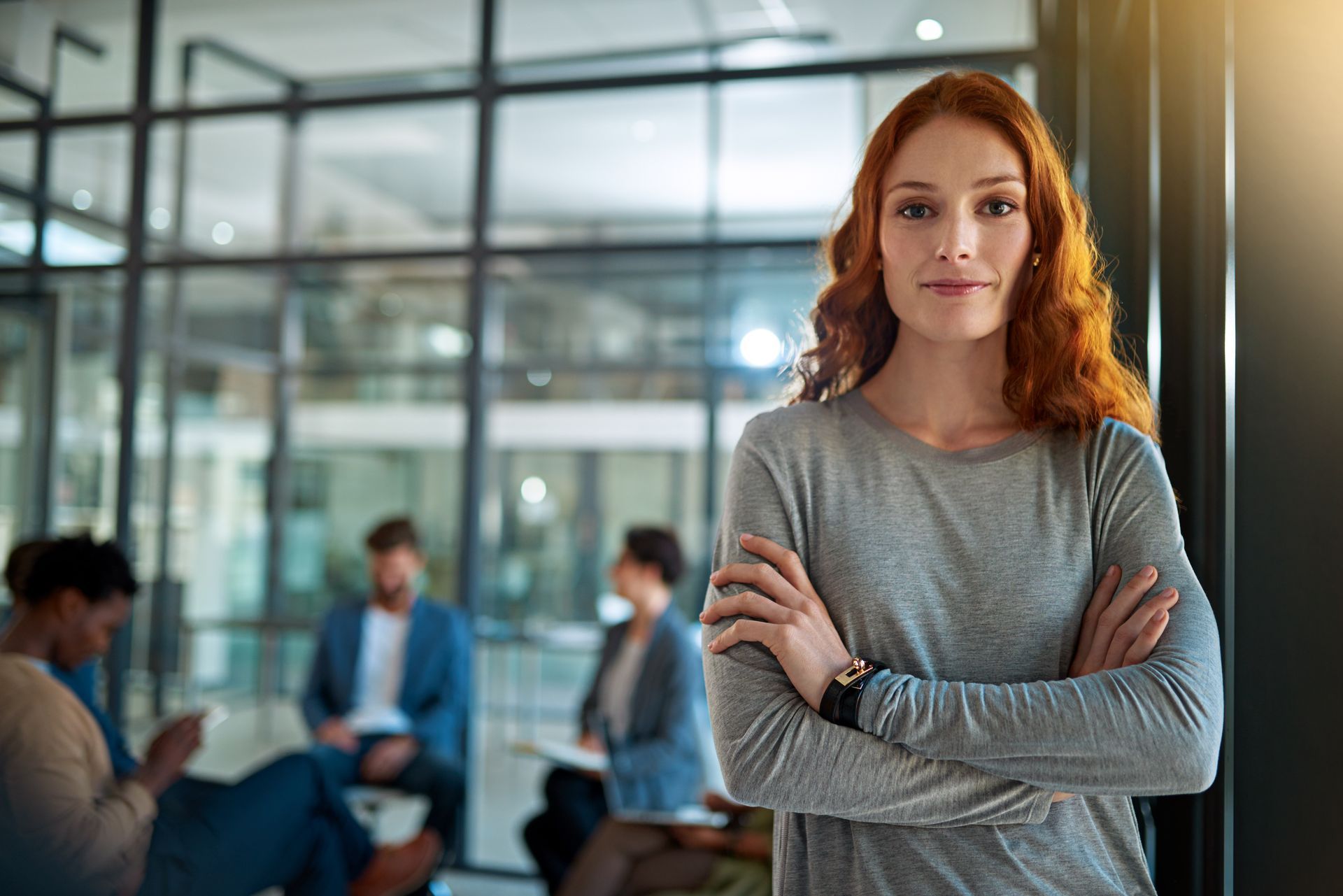 Woman with crossed arms, looking at camera in office with people in the background.