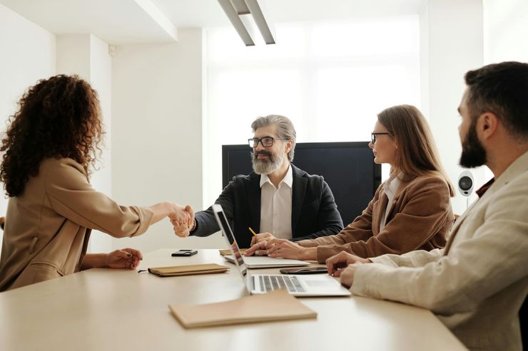 People in business attire shaking hands at a table, laptops present.