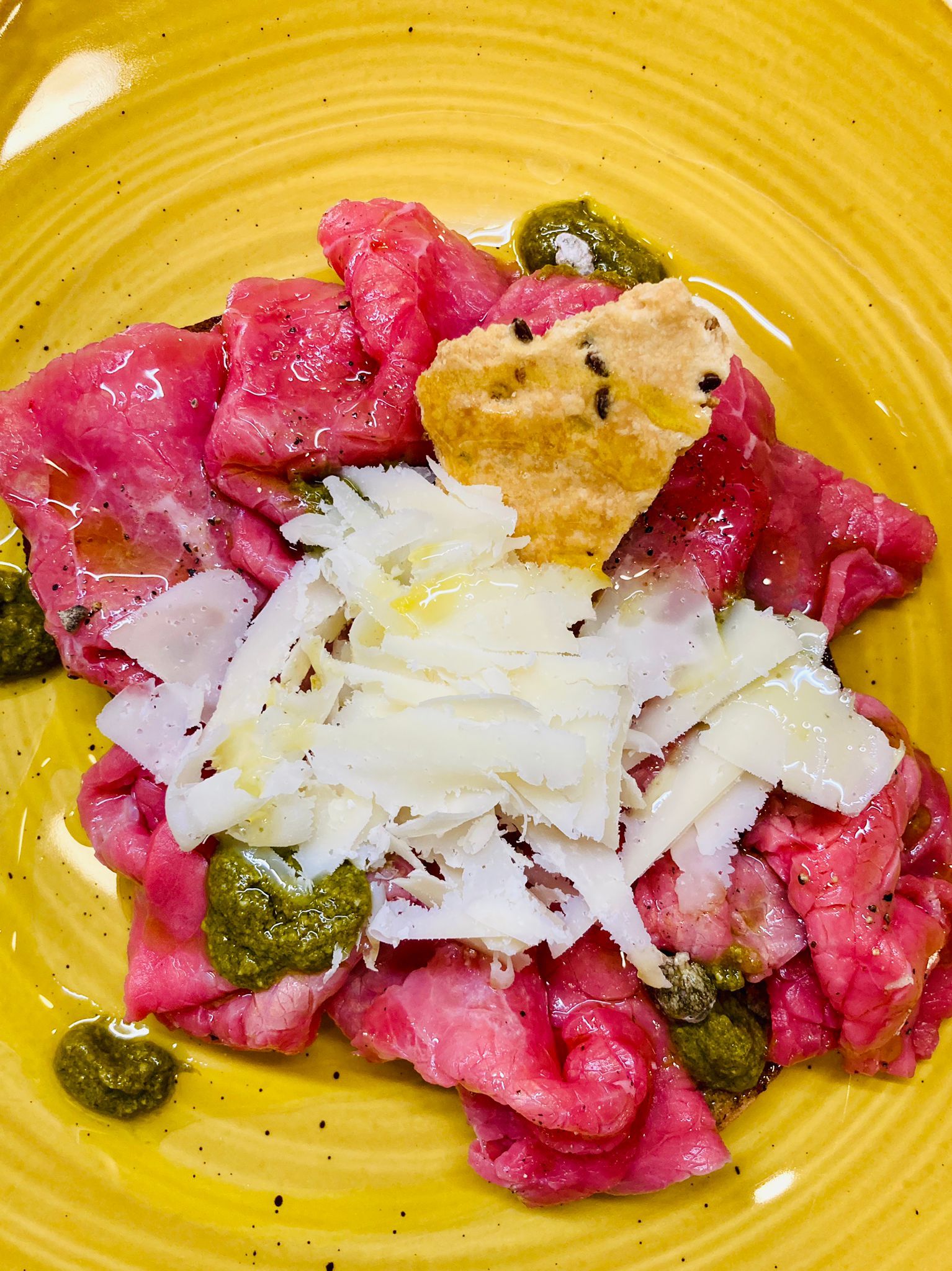 Carpaccio su un piatto giallo, guarnito con formaggio a scaglie, pesto e un cracker.