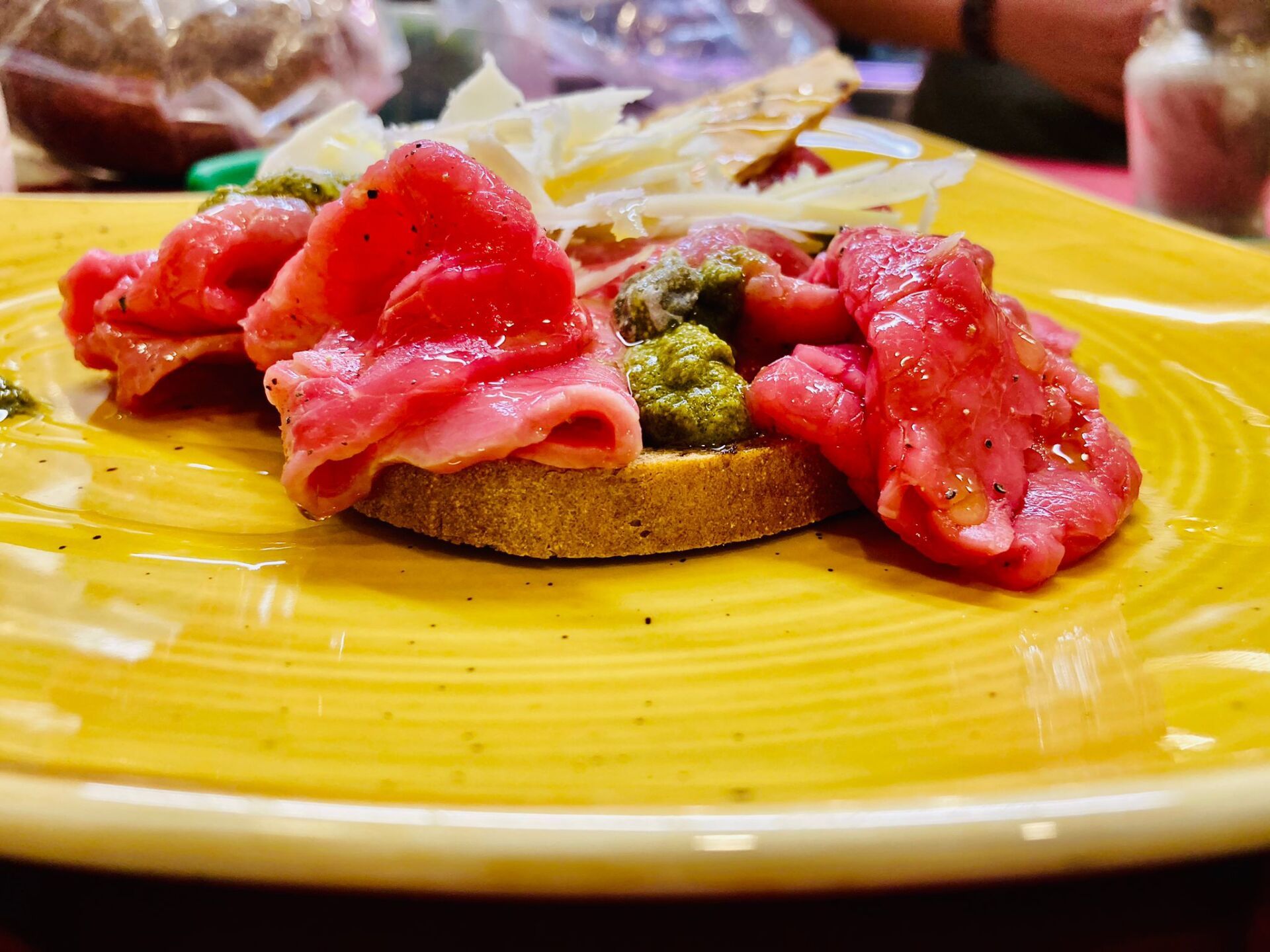Antipasto di carpaccio su un piatto giallo: manzo crudo tagliato sottile, pesto, formaggio e pane.