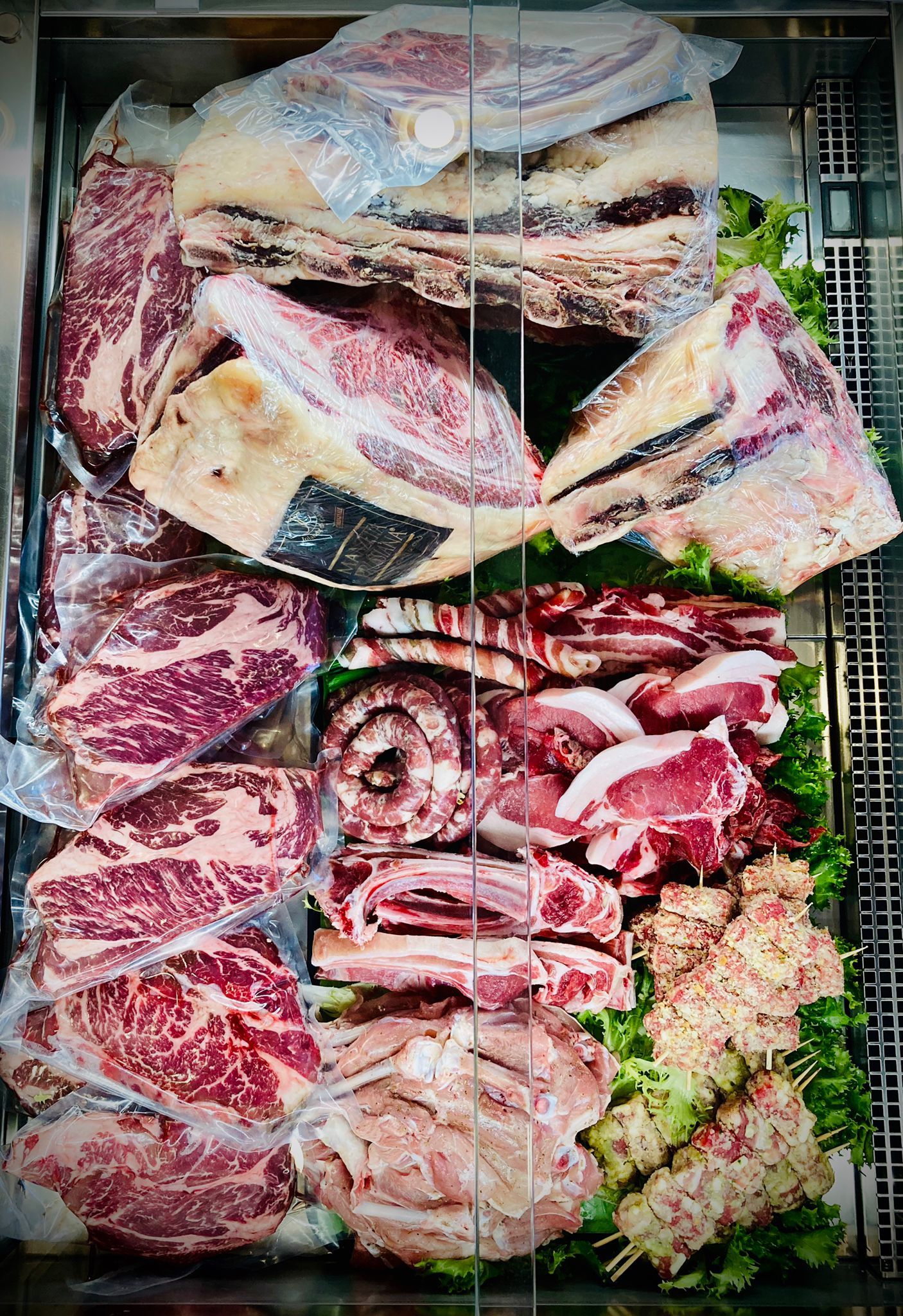 Vetrina con vari tagli di carne cruda, tra cui bistecche e spiedini, su un letto di verdure.