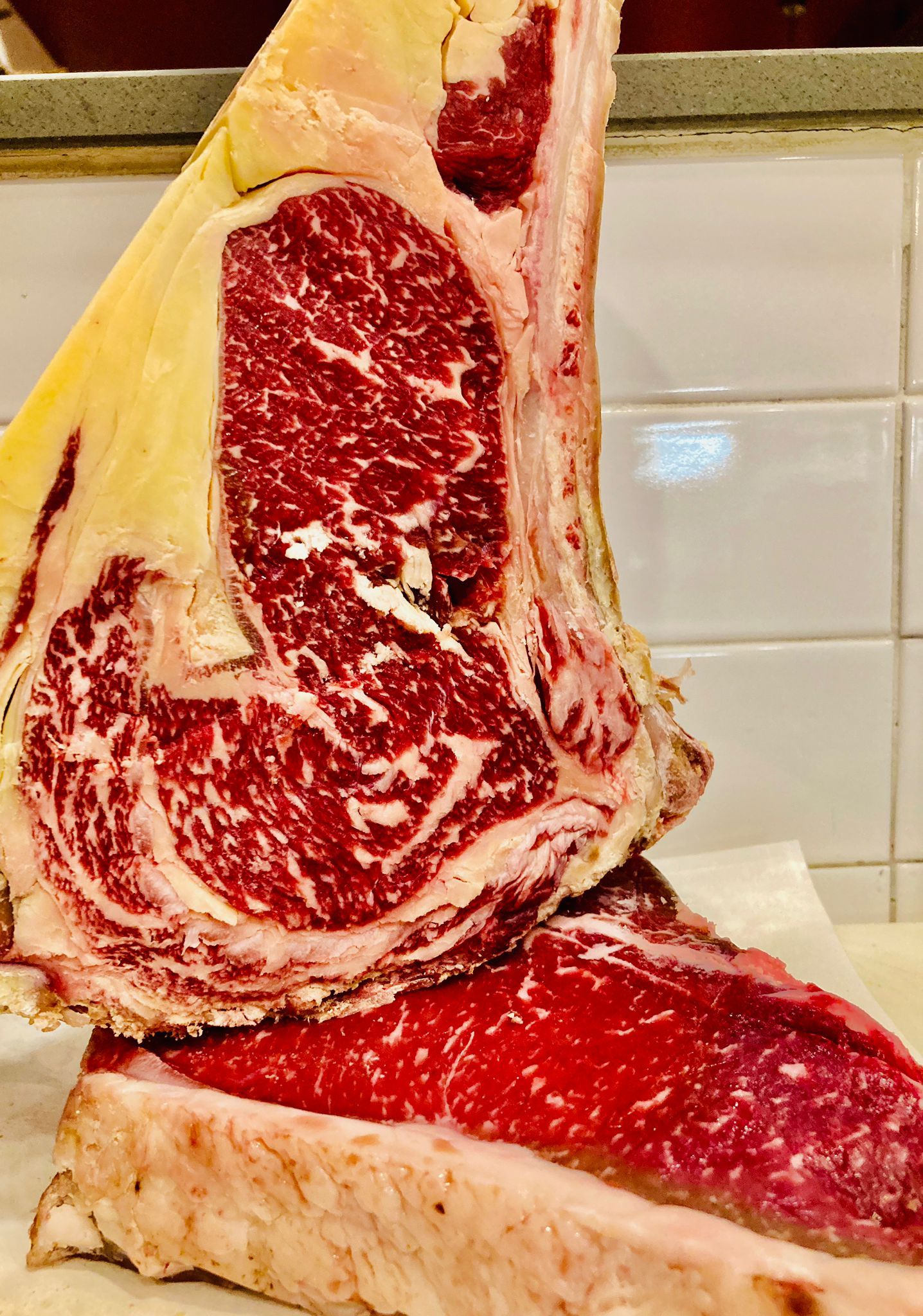 Primo piano di una grande bistecca cruda marmorizzata con grasso visibile e un osso su uno sfondo piastrellato.