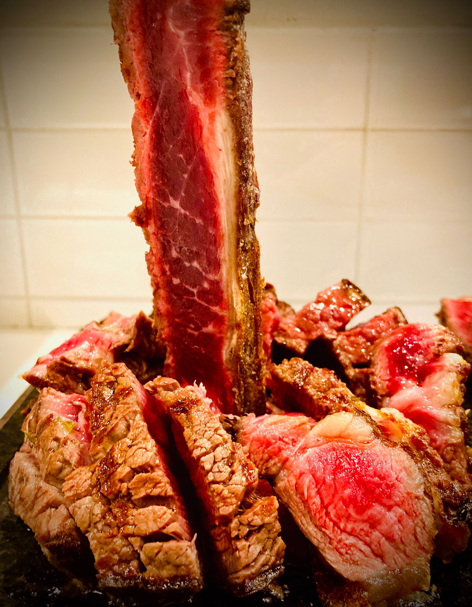 Bistecca tagliata a fette e condita su un piatto nero, con una grande striscia di carne verticale al centro, appoggiata contro una parete piastrellata.