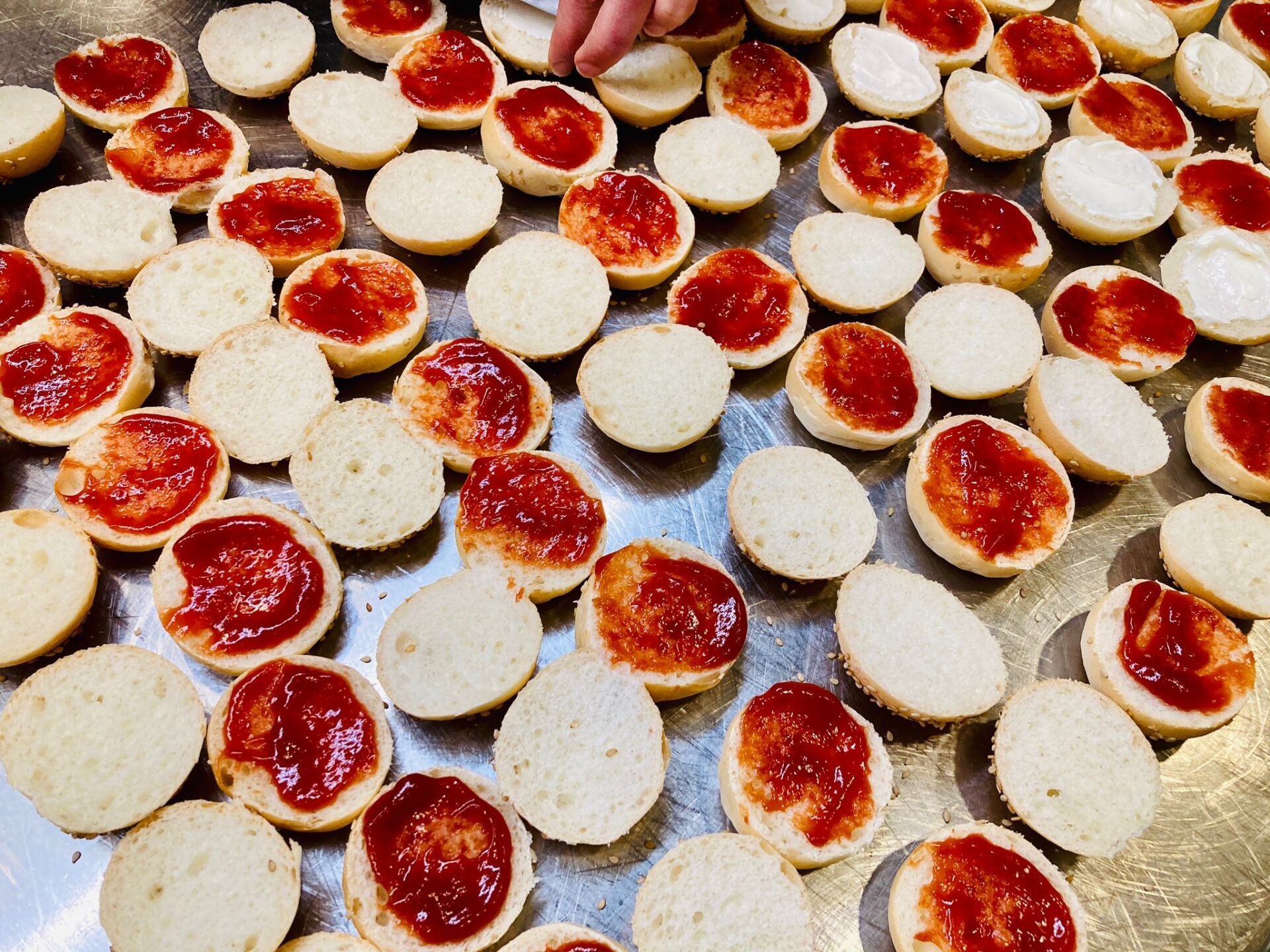 Metà di pane rotondo, alcune ricoperte di marmellata rossa, disposte su una superficie metallica.