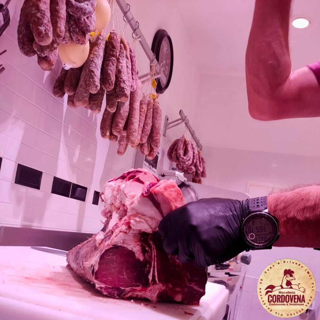 Un macellaio che affetta un grosso pezzo di carne cruda con salumi appesi sopra.
