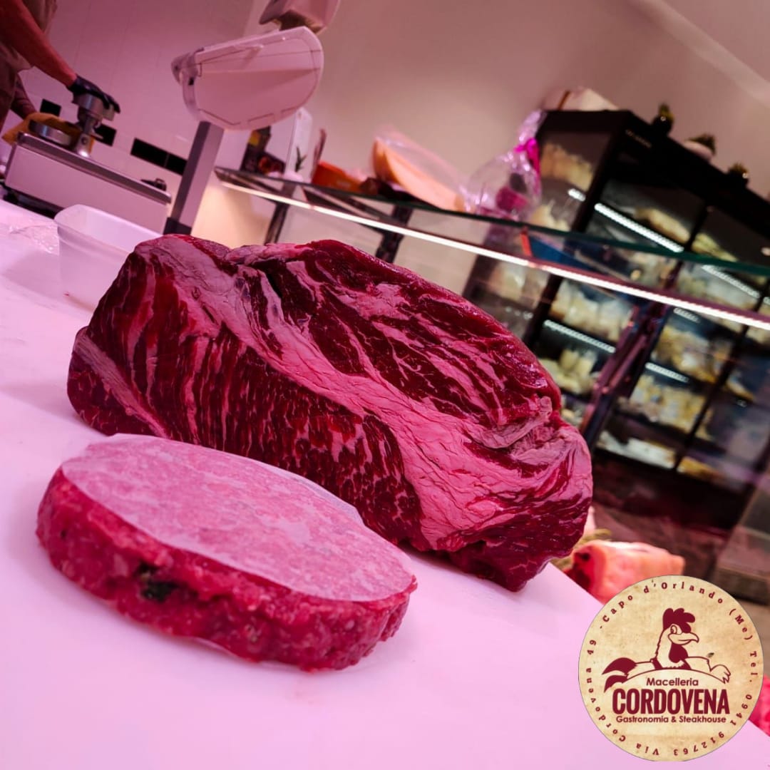 Tagli di carne cruda esposti sul bancone di una macelleria, tra cui una grande bistecca e un hamburger.