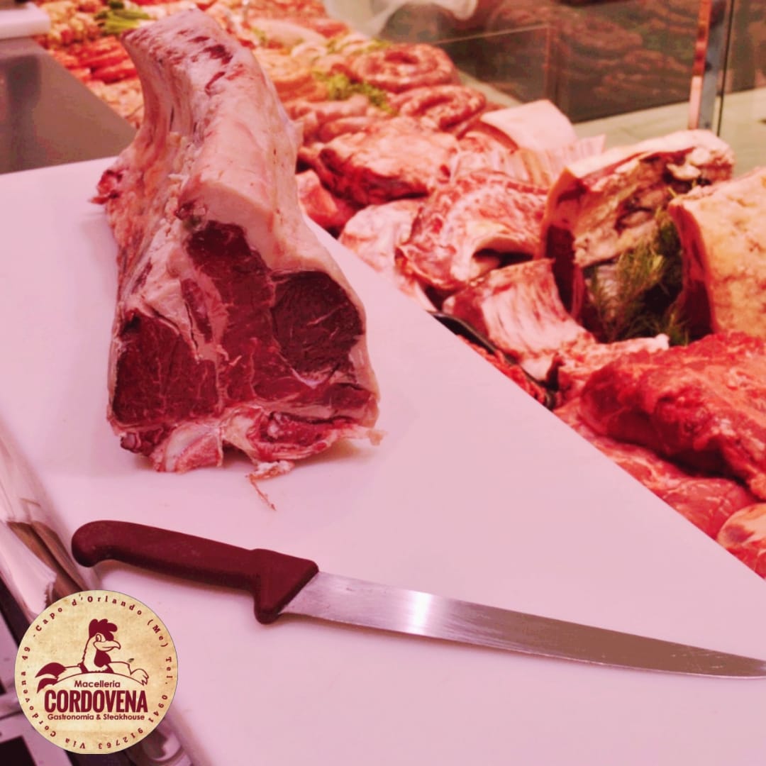 Un grosso taglio di carne cruda è appoggiato su un tagliere bianco, accanto a un coltello. Altri tagli di carne sono sullo sfondo.