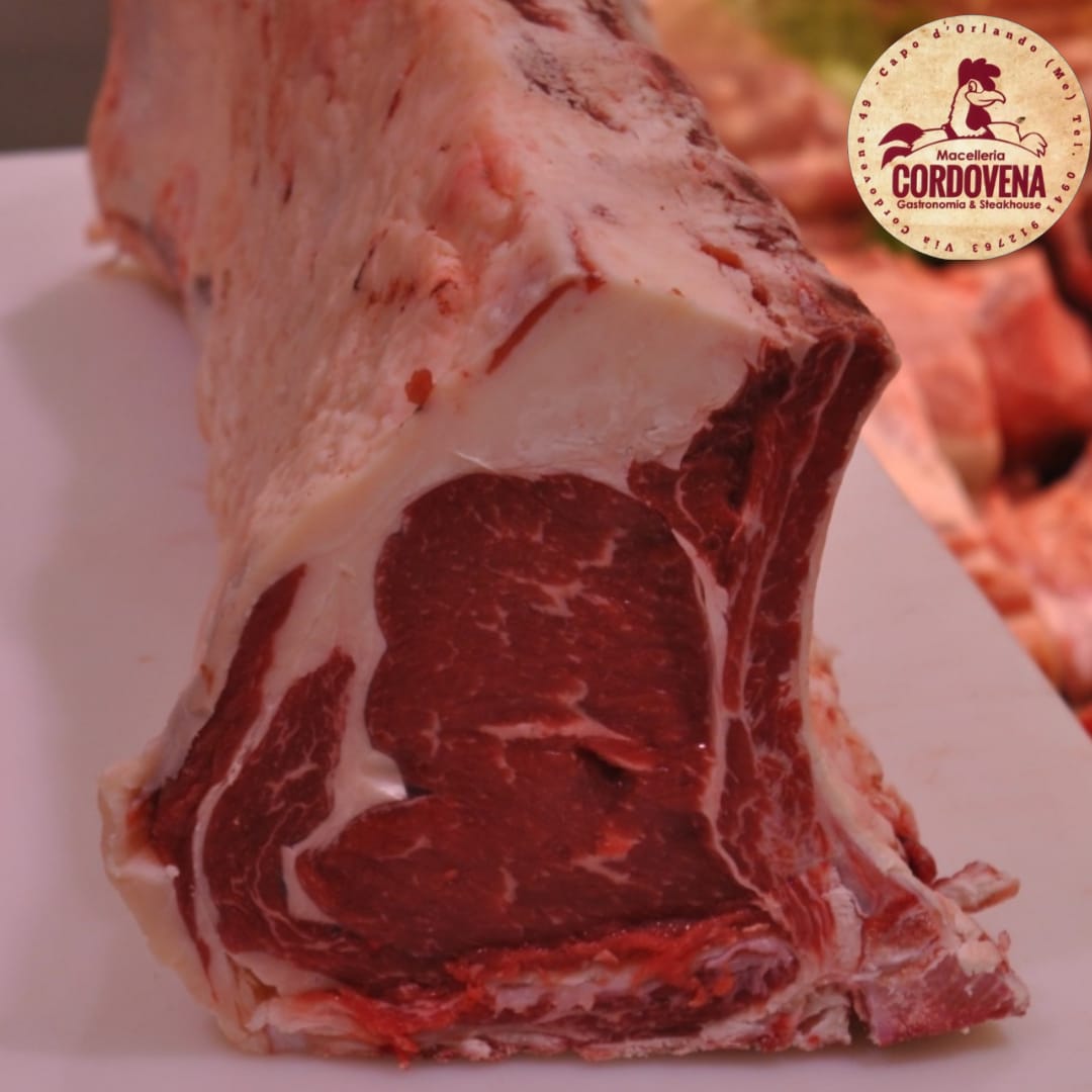 Bistecca di ribeye cruda, tagliata spessa, con marezzature e osso visibili, su una superficie bianca.