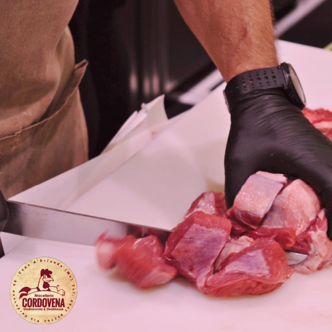 Persona con guanti neri che taglia carne cruda su una superficie bianca. Logo 