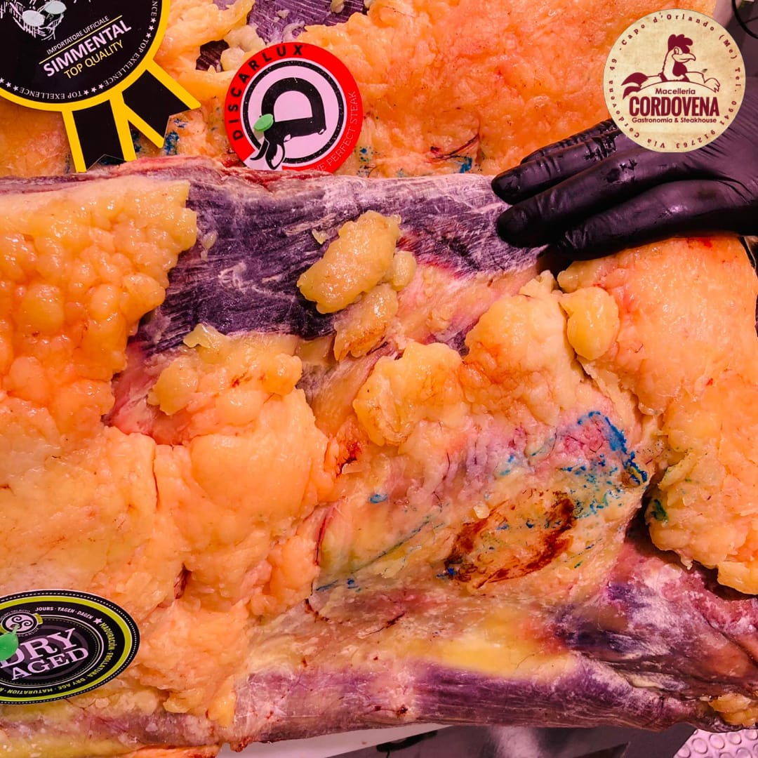 Primo piano di carne cruda con grasso giallo e una mano guantata di nero. Sono visibili etichette e loghi.