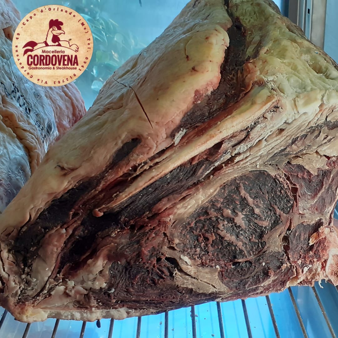 Grande arrosto di manzo frollato a secco, dalla superficie scura e screziata, adagiato su una griglia metallica bluastra.