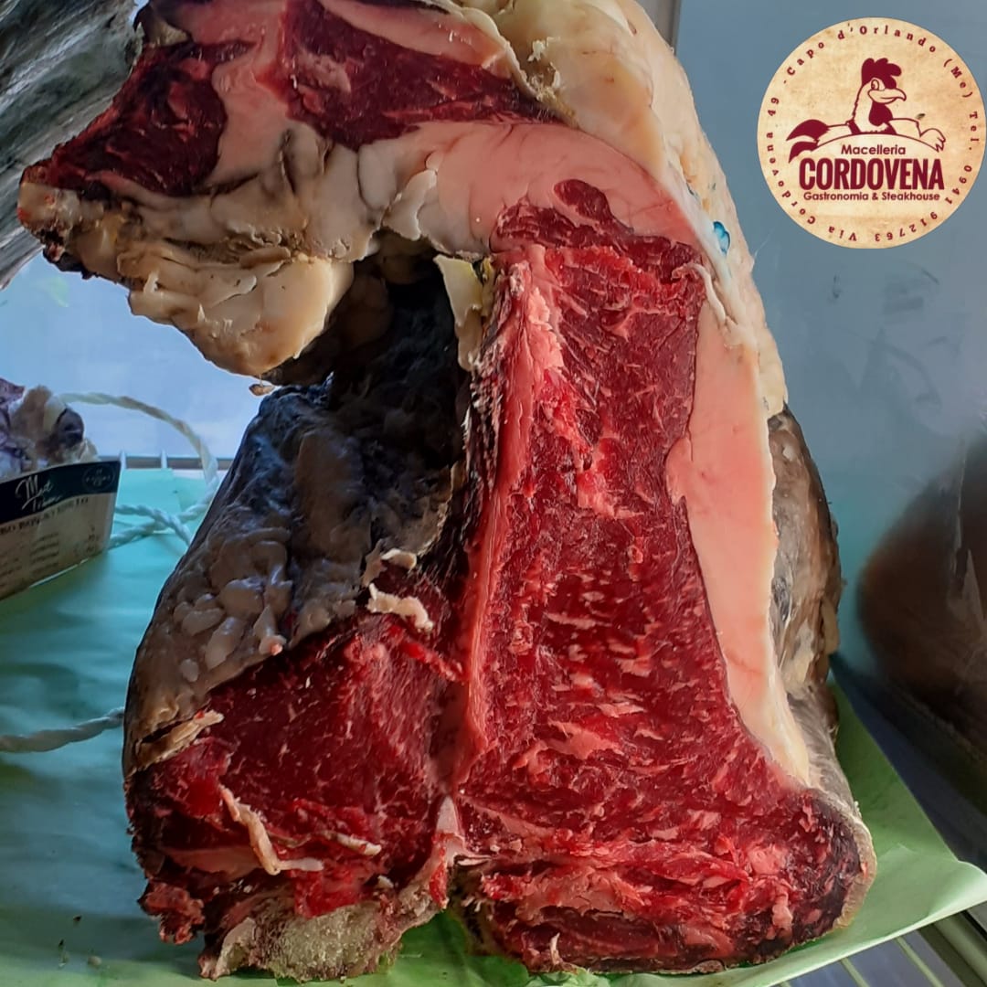 Grande bistecca cruda, carne rossa con marezzature bianche, su una superficie verde. Logo Cordoveña in un angolo.