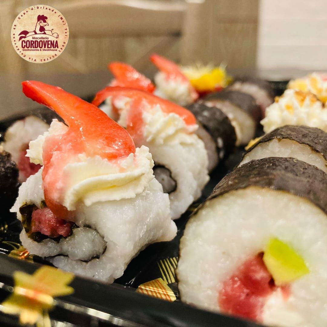 Involtini di sushi serviti su un vassoio nero, guarniti con gamberi e panna, accanto ad altri con pesce e avocado.
