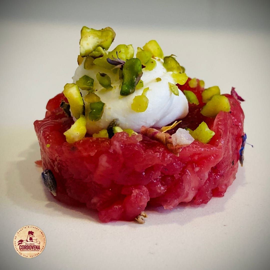 Tartare rossa guarnita con crema bianca e pistacchi verdi su una superficie bianca.