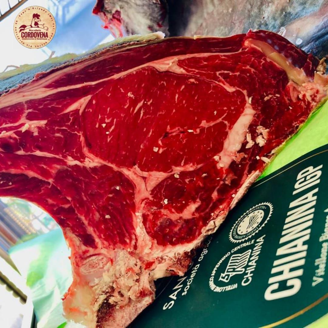 Bistecca di Chianina IGP cruda, tagliata spessa, con osso, carne rossa con marezzature di grasso, su superficie verde.