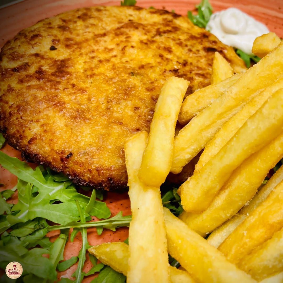 Cotoletta impanata con patatine fritte e un cucchiaio di salsa su un letto di verdure, servita su un piatto arancione.