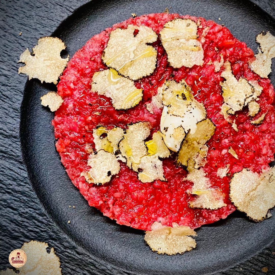 Tartare di manzo guarnita con fettine di tartufo su un piatto nero.