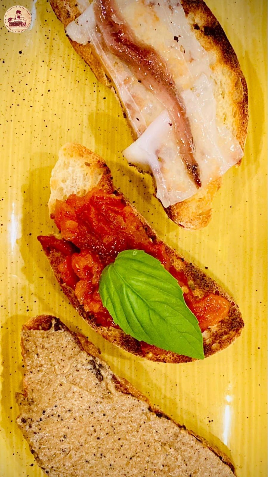 Pane tostato condito con pomodoro, basilico, acciughe e paté, disposto su un piatto giallo.