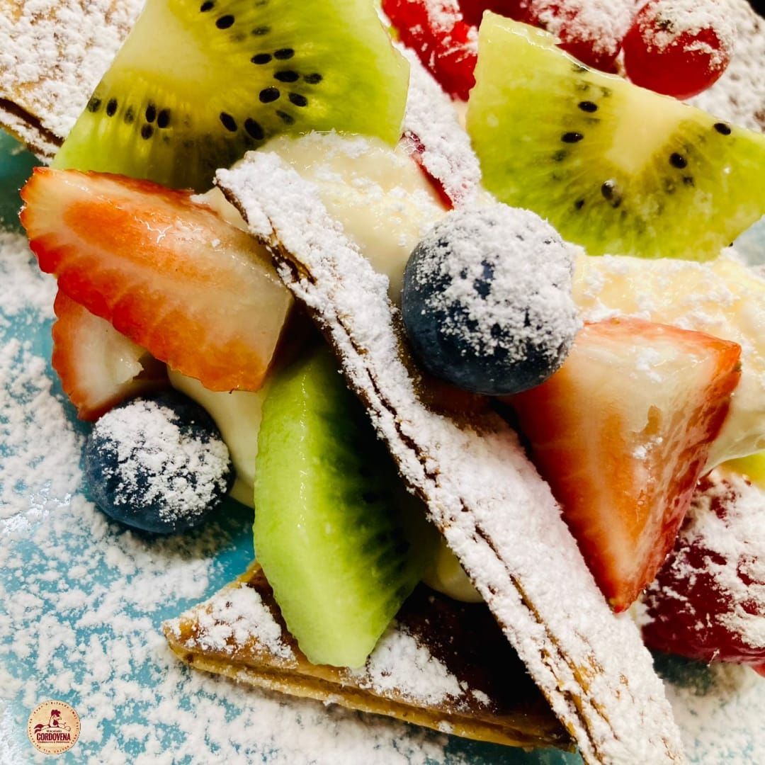 Primo piano di un dessert con kiwi, fragole, mirtilli e zucchero a velo su un piatto blu.