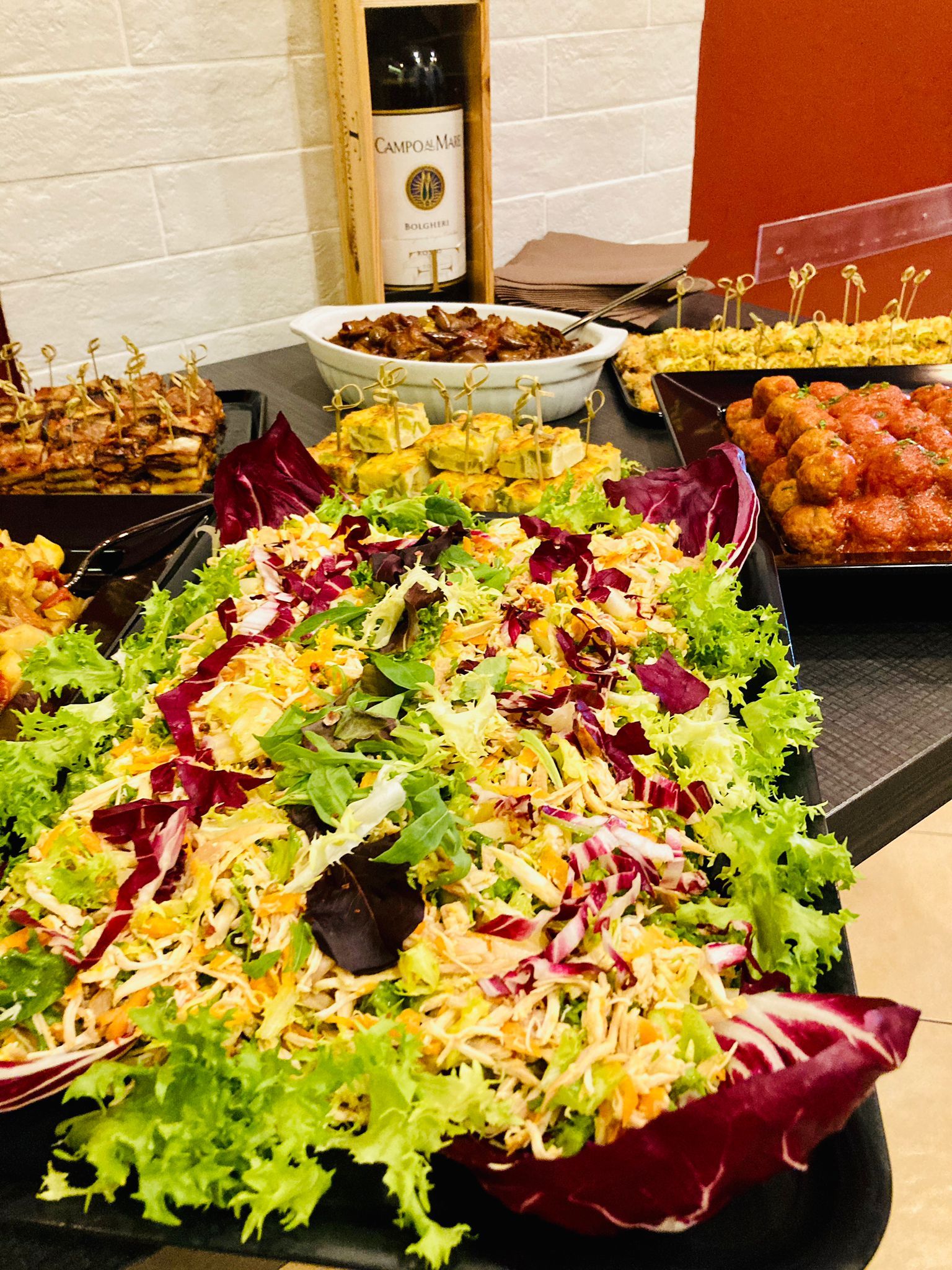 Buffet con insalata, polpette, spiedini e bottiglia di vino su un tavolo.