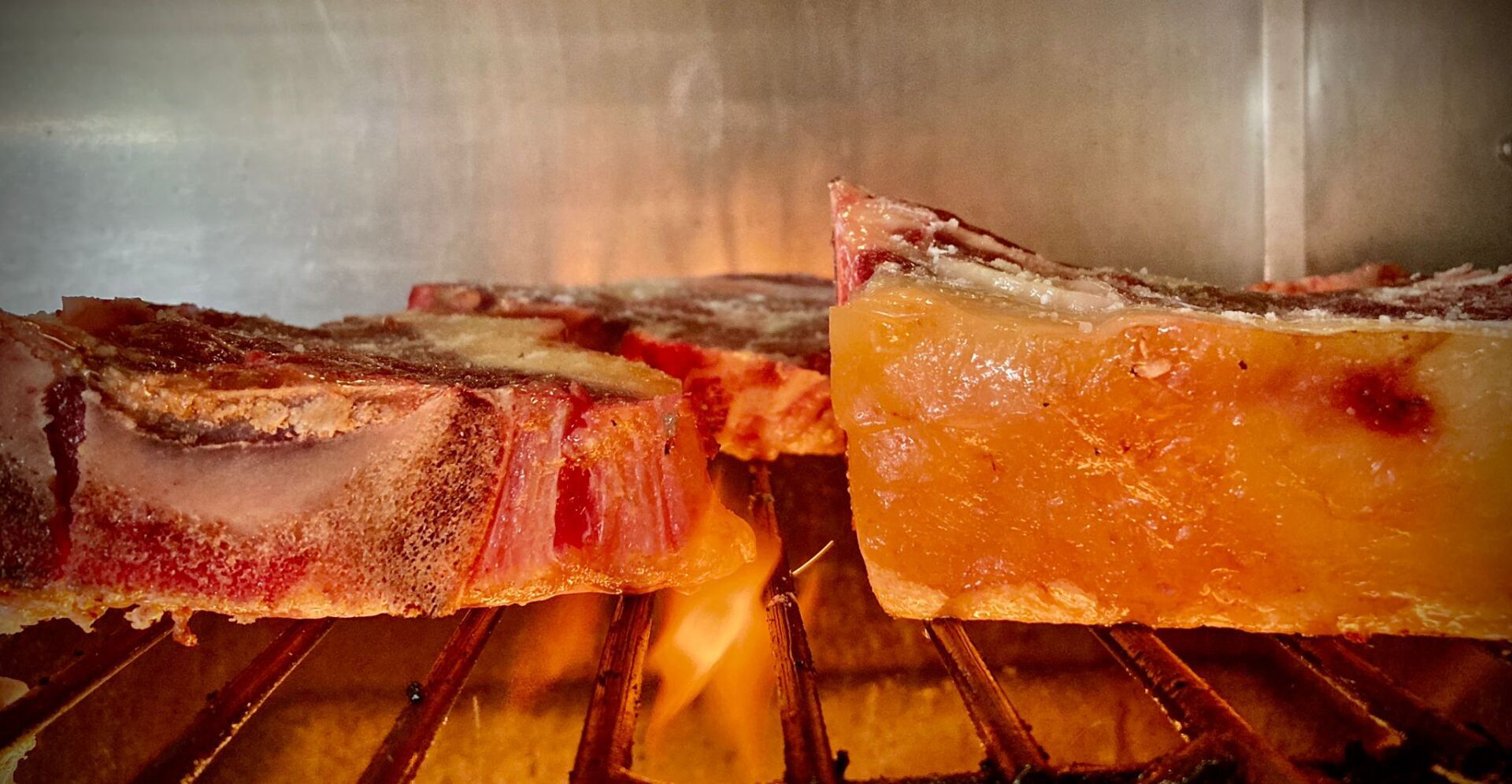 Primo piano di bistecche tagliate spesse, cotte alla griglia su una griglia sopra le fiamme, che mettono in mostra i bordi bruciacchiati e l'interno succoso.