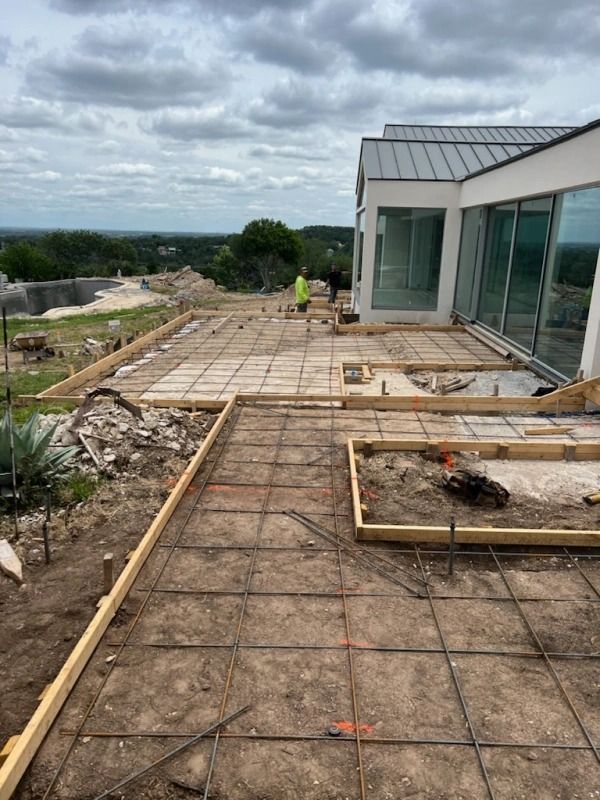 Construction site with wooden forms, rebar grid, and a modern house; cloudy sky.