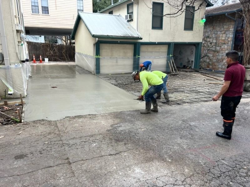 Construction workers pouring concrete on a driveway, near a garage. One worker watches.