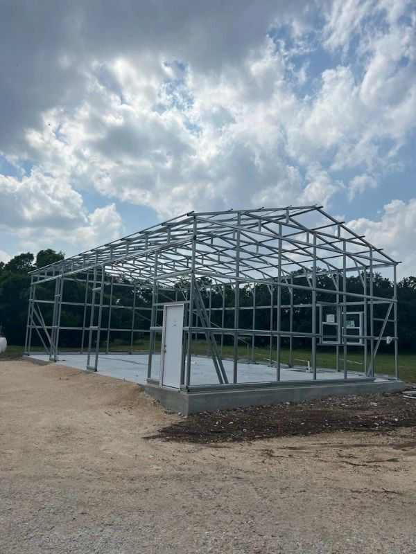 Steel-framed building under construction against a cloudy sky. Framing includes door and window openings on a concrete slab.
