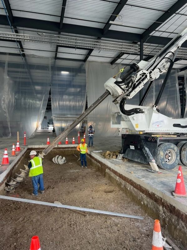 Workers use a drilling machine in a building's excavation. Construction workers in safety vests, orange cones.