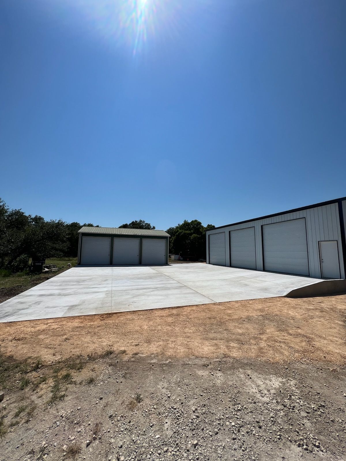 Two metal buildings with large doors on a concrete pad under a clear blue sky.
