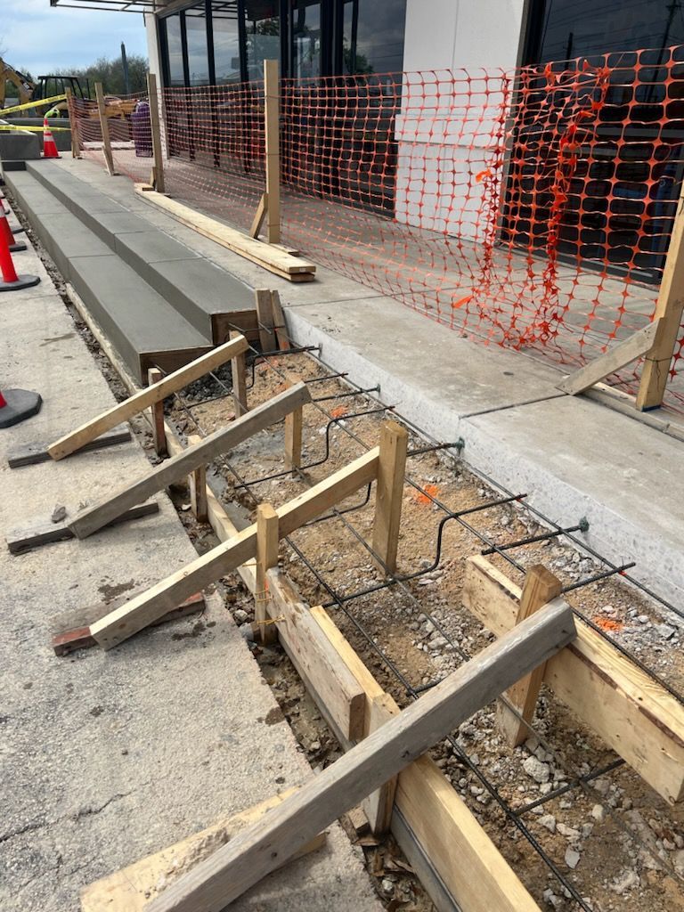 Construction site: Wooden forms, rebar, and poured concrete sidewalk along a building. Orange safety fence.