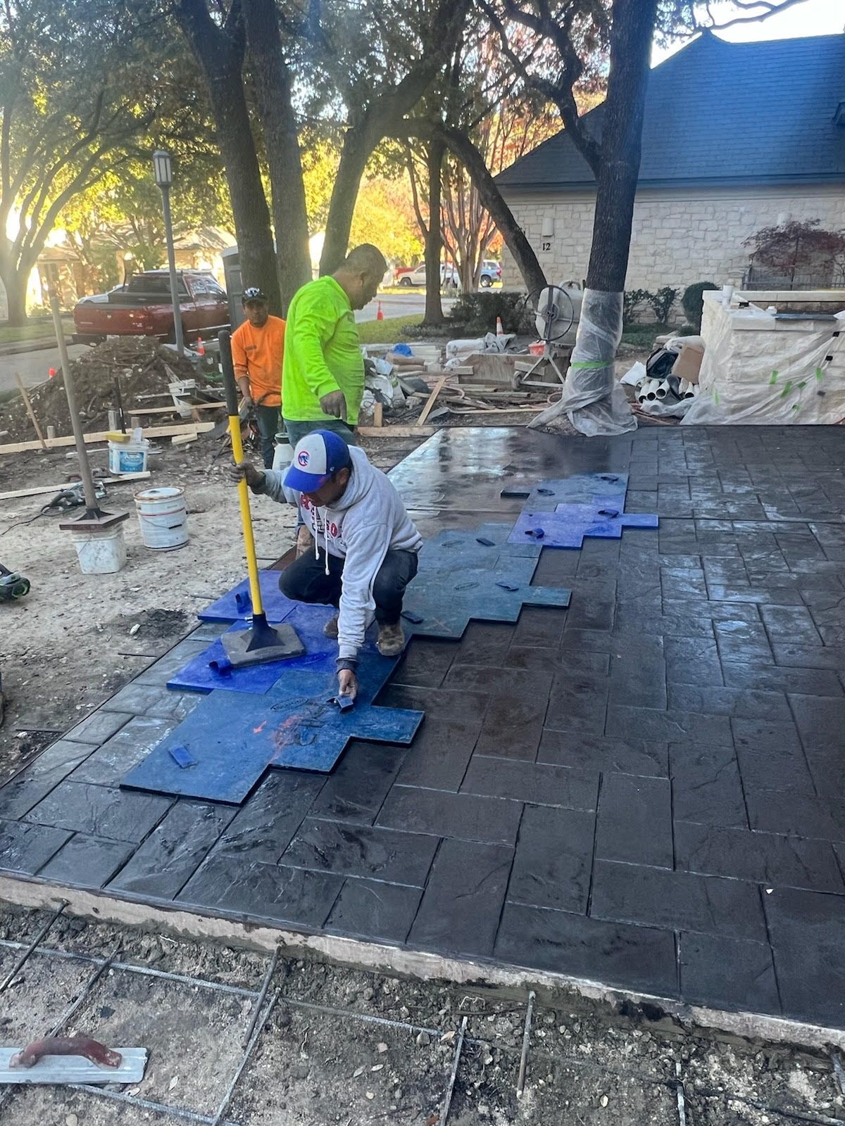 Workers stamping wet concrete, creating a patterned patio. Blue stamps, bright clothing, outdoor setting.