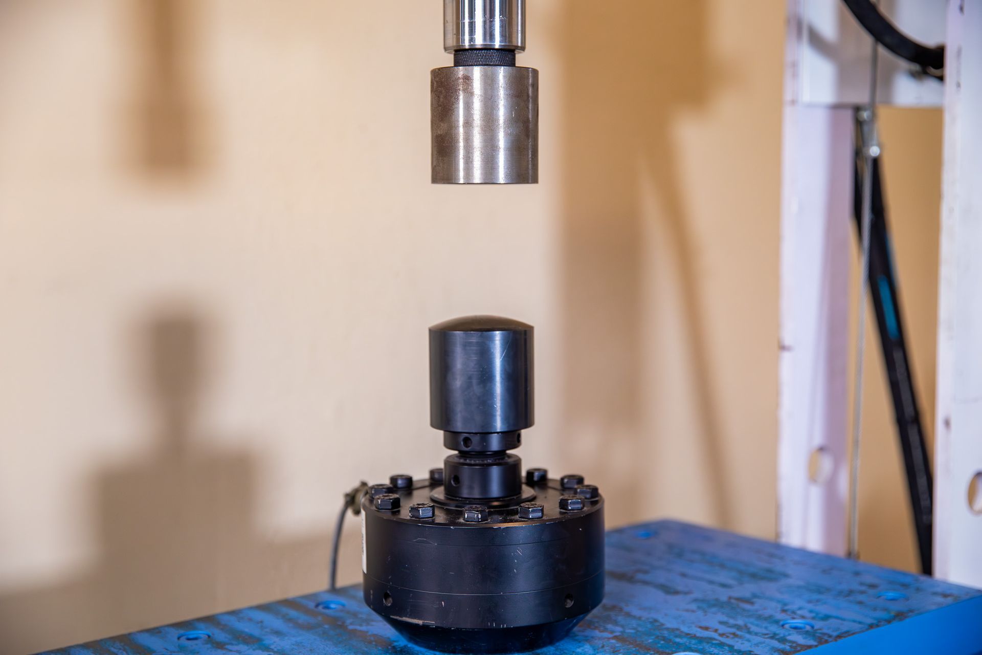 A hydraulic press over a black metal object with a round top on a blue surface.