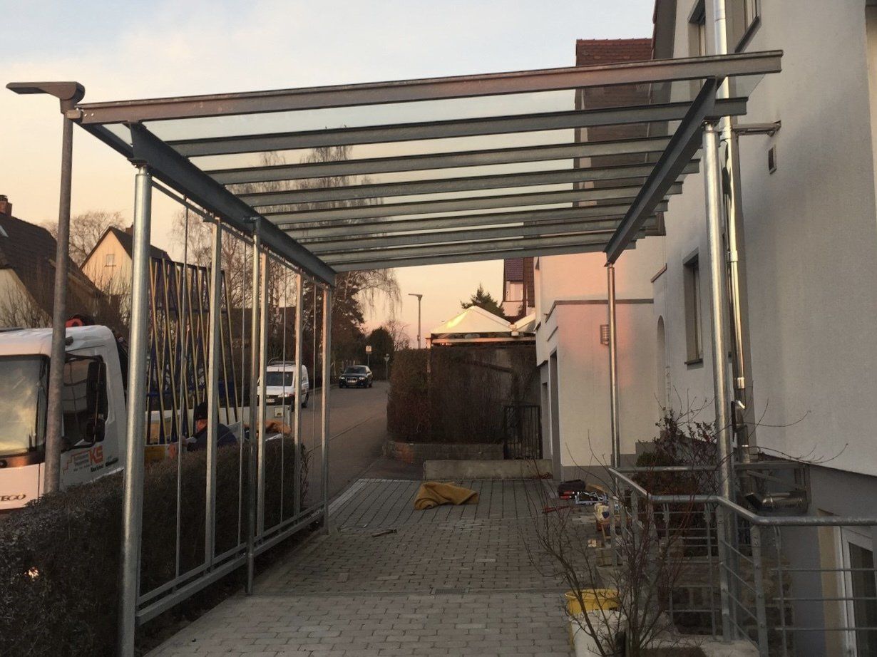 Vor einem weißen Gebäude entsteht ein Carport mit Glasdach.