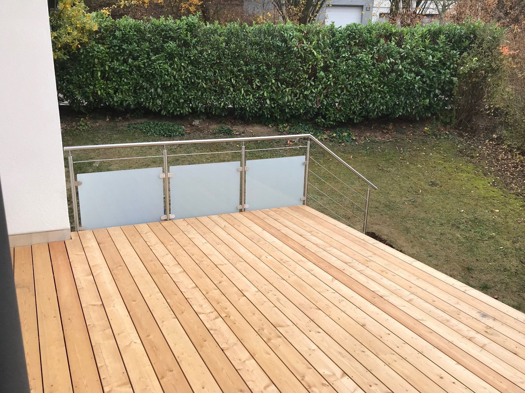 Ein Holzdeck mit einem Edelstahlgeländer und einer Hecke im Hintergrund.