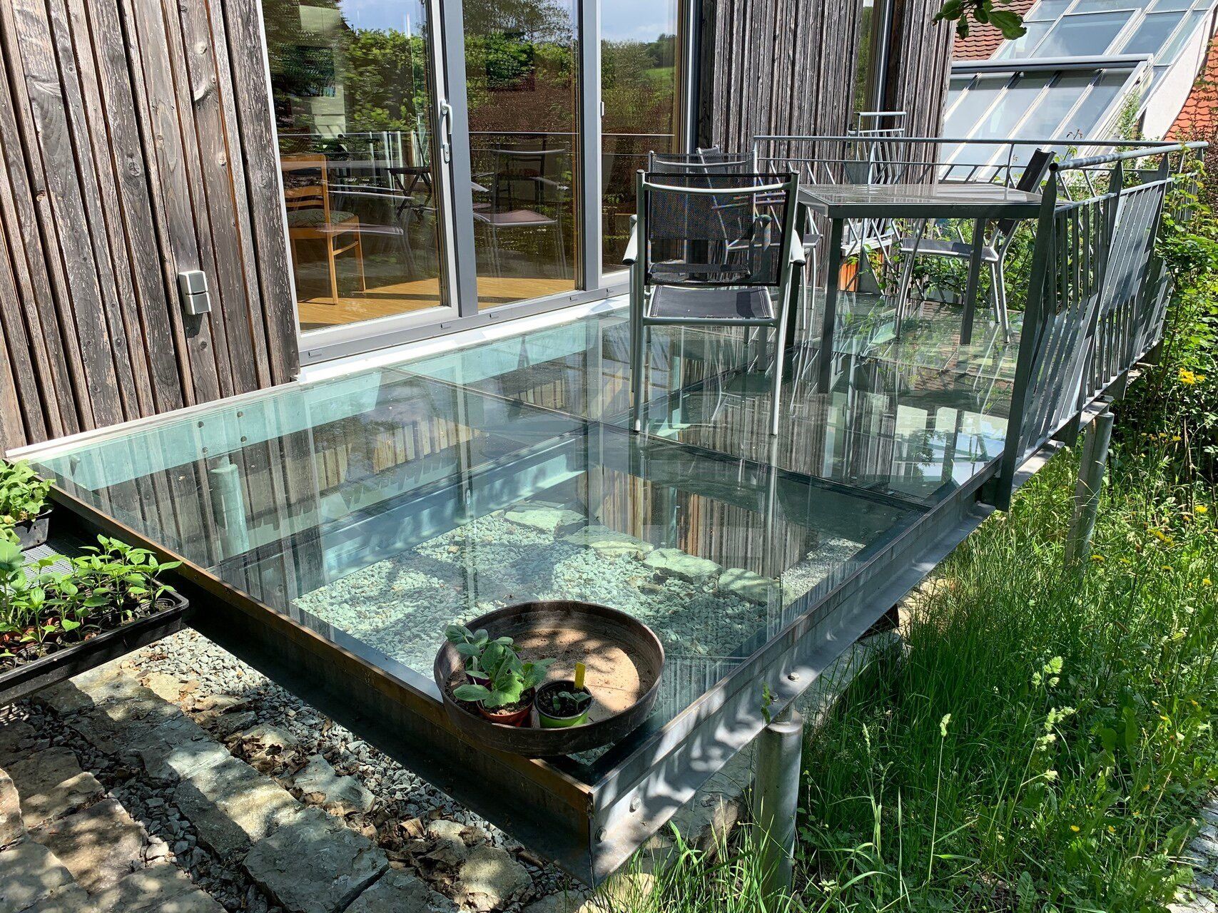 Auf einer Terrasse neben einem Haus steht ein Glastisch mit einer Schale voller Pflanzen darauf.